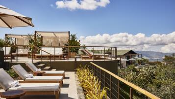 A rooftop lounge area with sun loungers, umbrellas, and a view of the mountains.