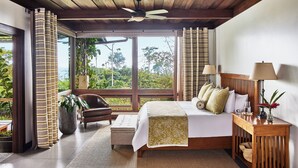 Un dormitorio amplio con una cama grande, cabecera de madera, mesita de noche, una silla y vistas al exterior a través de amplias ventanas.