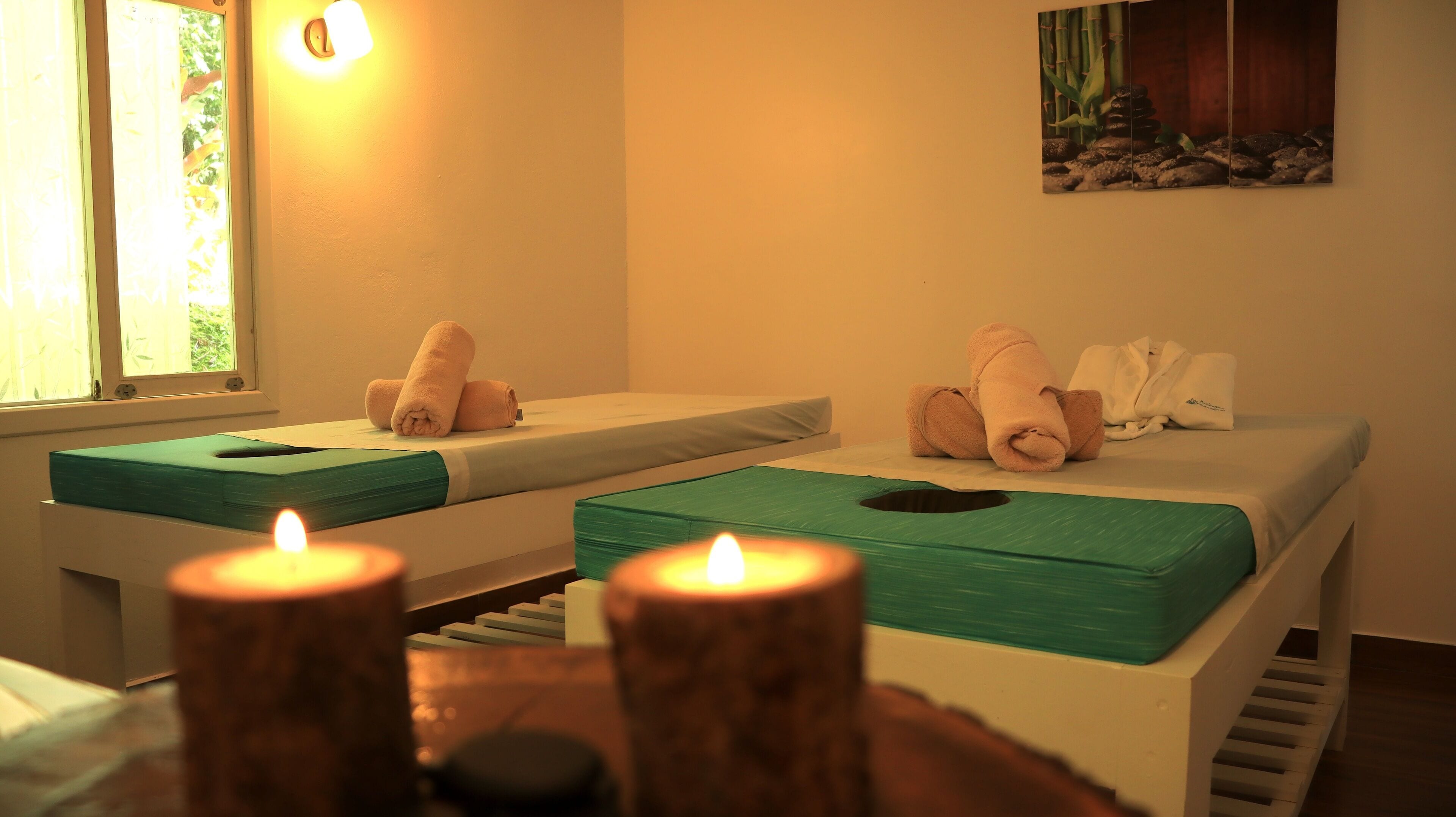 A spa room with two massage tables, each with a green mat and towels. There are candles on a wooden table in the foreground.
