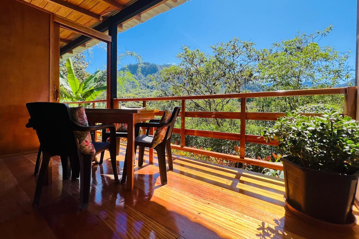 A wooden deck with a table and chairs, overlooking a lush landscape with trees and mountains.