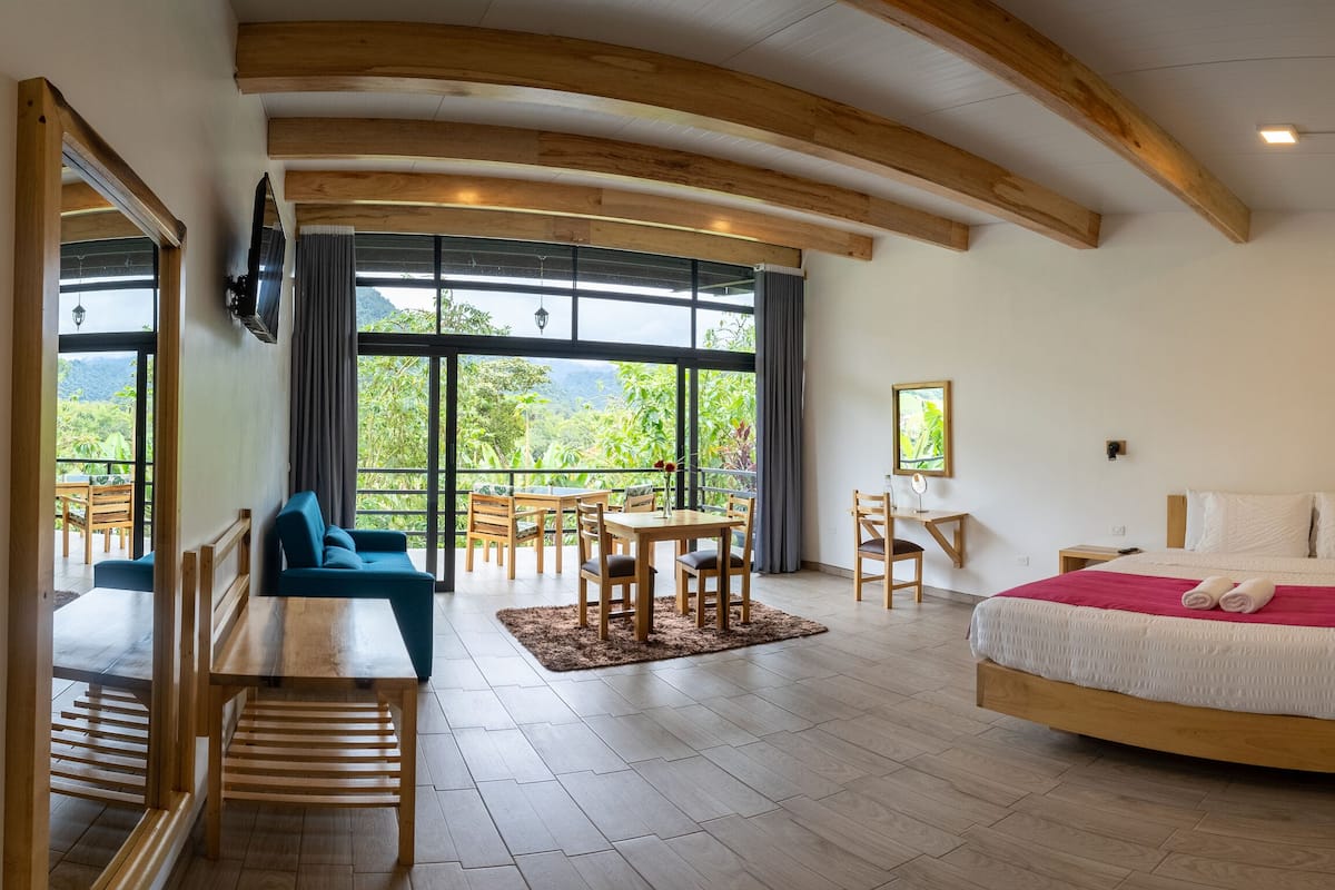 A spacious bedroom with a large bed, wooden ceiling beams, a dining area with a table and chairs, and a view of greenery through large windows.