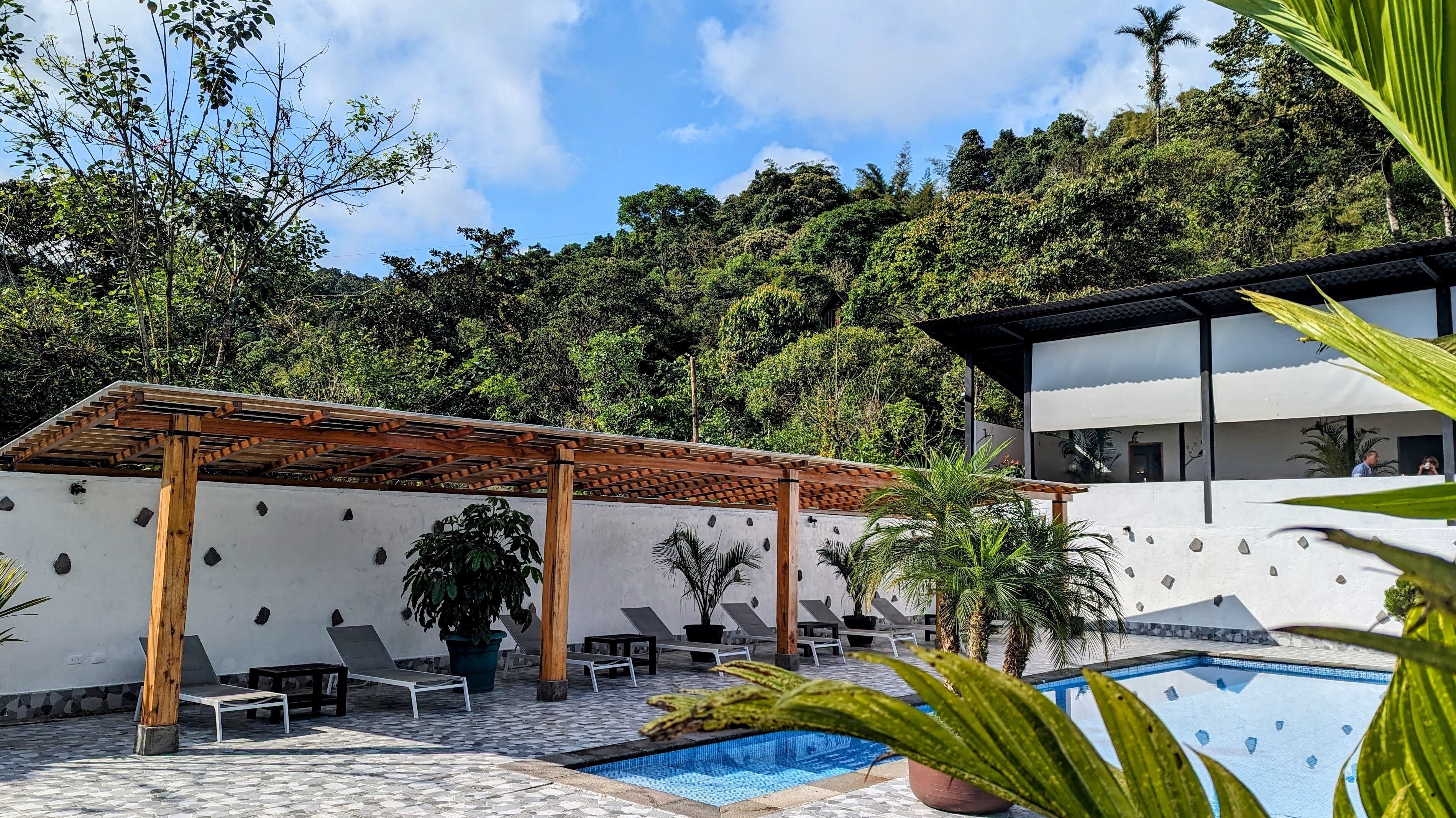 Zona de piscina con sillones de descanso, una pérgola y un edificio moderno de color blanco.