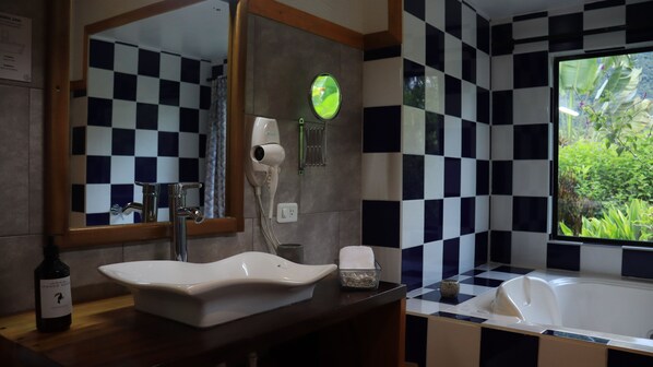 Un baño con salpicadero de azulejos a cuadros, un lavamanos, un espejo, un secador de pelo y una ventana con vistas al verdor.