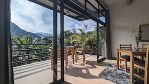 Un balcón con vistas a montañas y vegetación tropical, muebles de madera y un ventanal de vidrio.