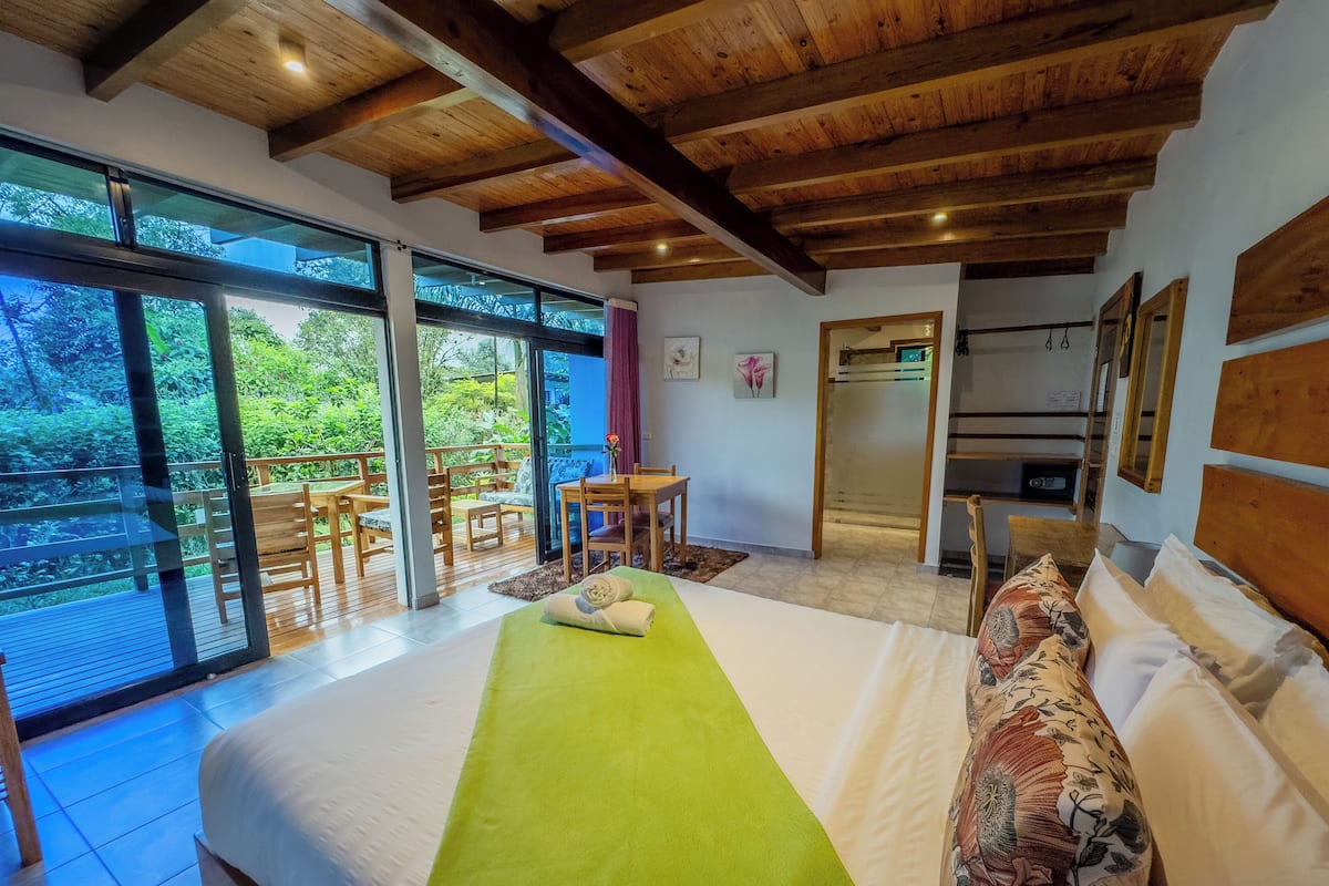 A bedroom with a large bed, a green bedspread, and a view of a balcony with wooden furniture.