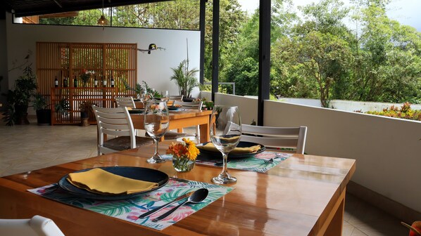 Un comedor con una mesa de madera dispuesta para dos, un pájaro decorativo en la pared y vistas a abundante vegetación.
