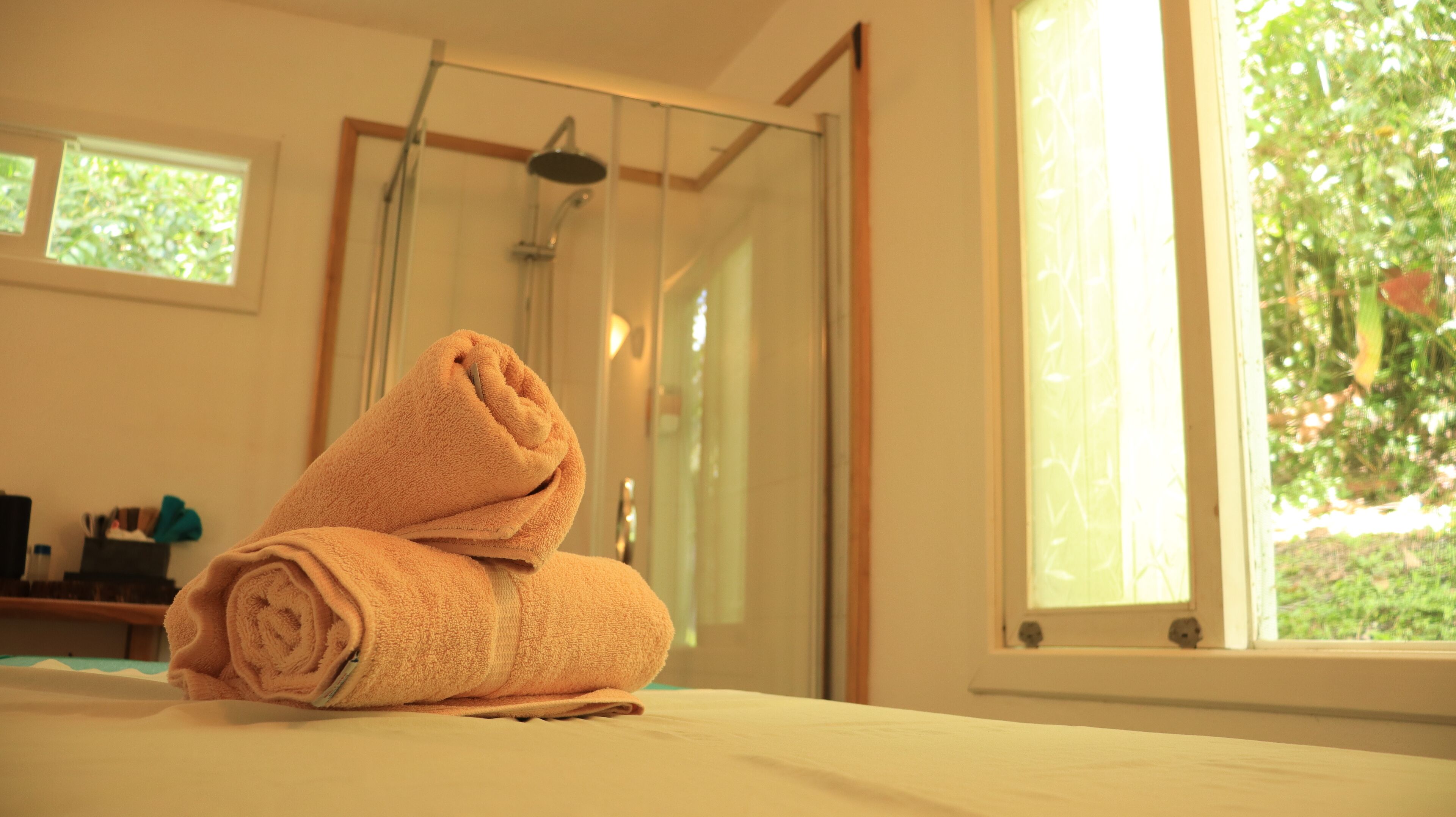 Una cama bien hecha, una toalla enrollada, una puerta de ducha de vidrio y una ventana con vistas a follaje.