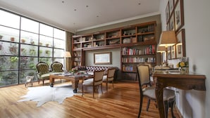 A spacious living room with a large window, wooden flooring, a leather sofa, a coffee table, and a bookshelf filled with books and decorative items. - Alma Histórica Boutique Hotel (Montevideo)
