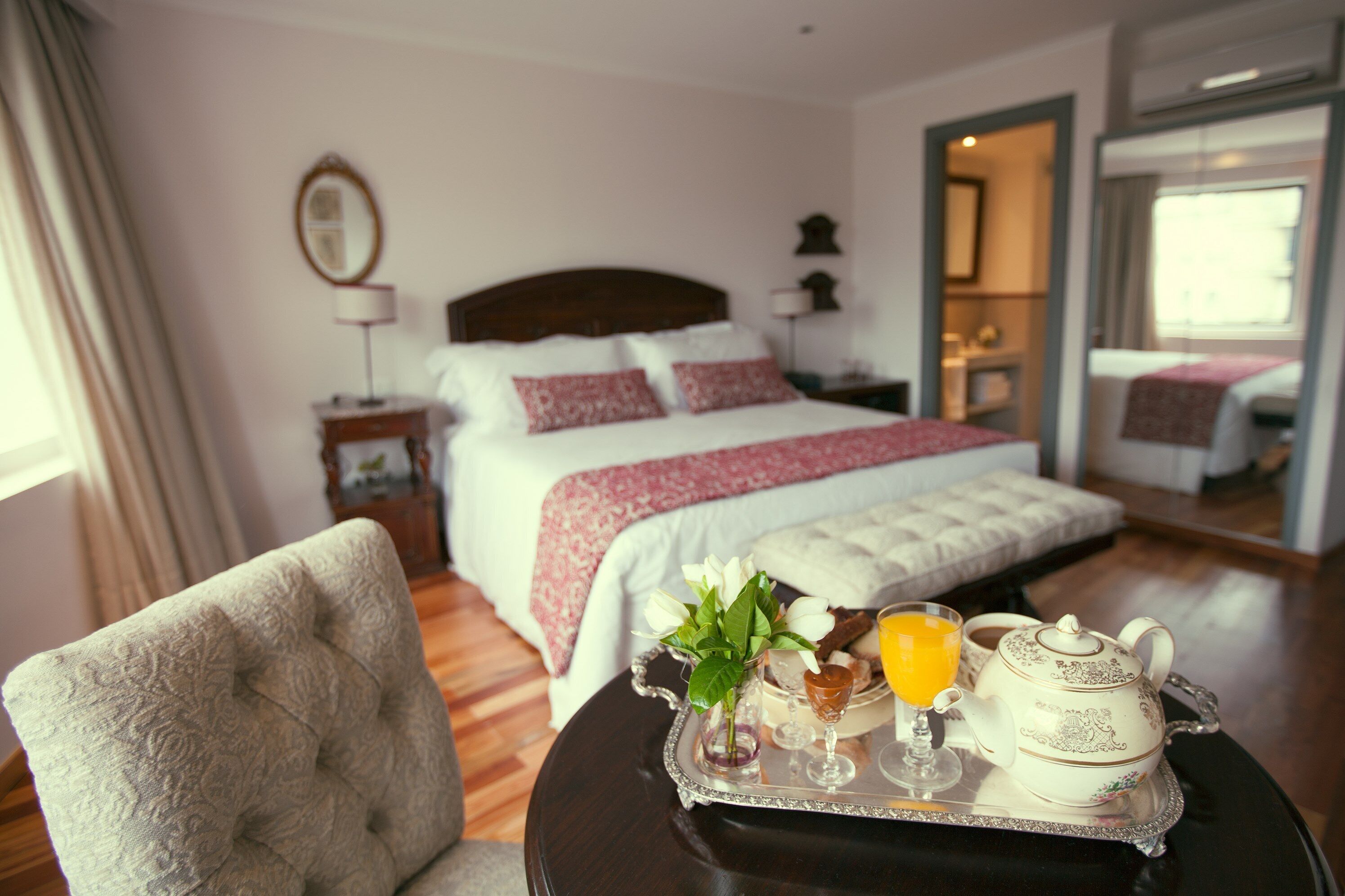 Une chambre d’hôtel avec un lit, une table de chevet, une chaise et un plateau sur lequel se trouvent une théière, un verre de jus d’orange et un vase avec des fleurs.