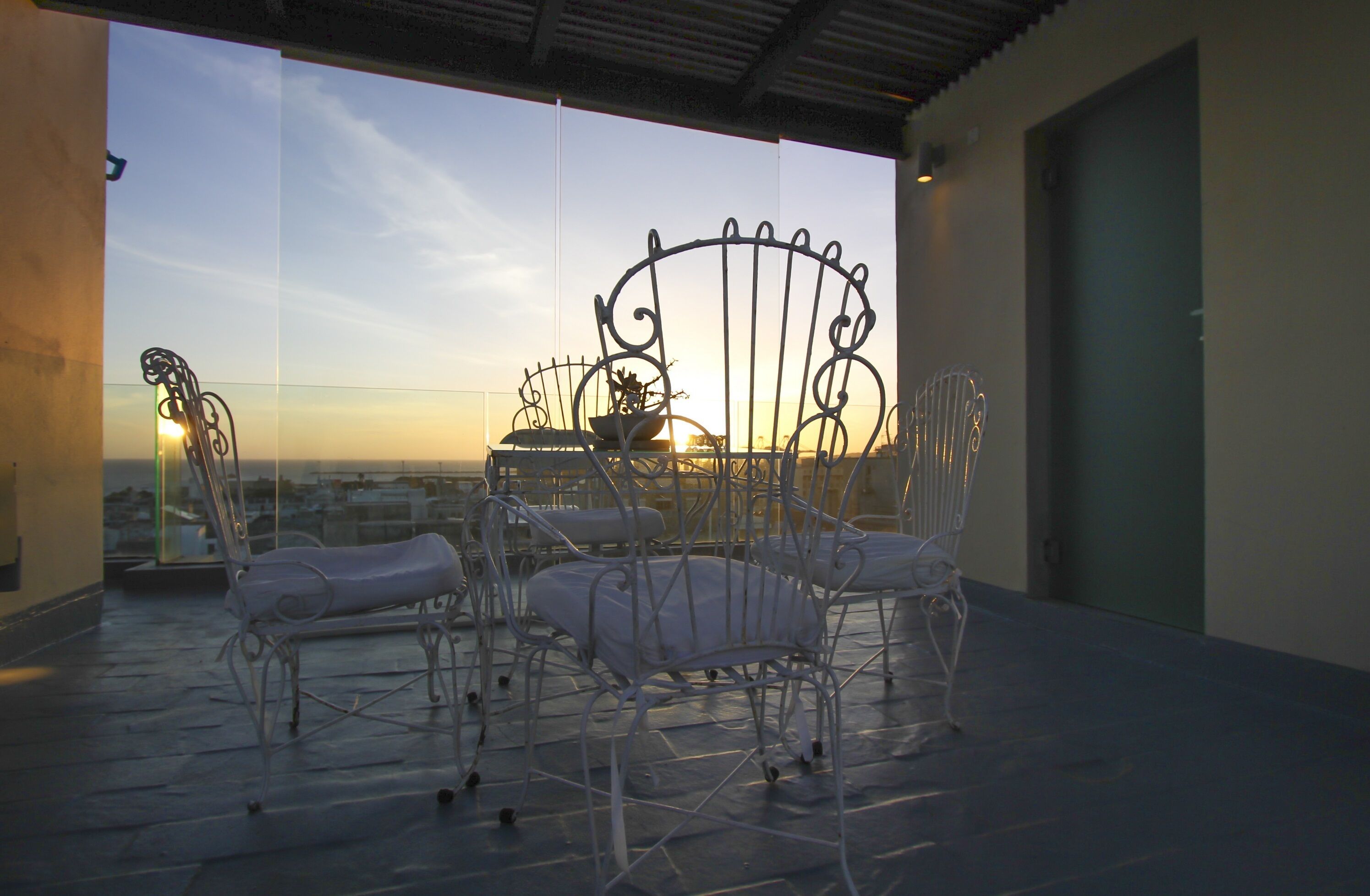 A rooftop terrace with metal chairs and a table, overlooking a cityscape at sunset.