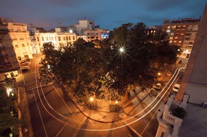 Un paisaje urbano nocturno con edificios iluminados, una calle bordeada de árboles y estelas de luz dejadas por vehículos en movimiento.