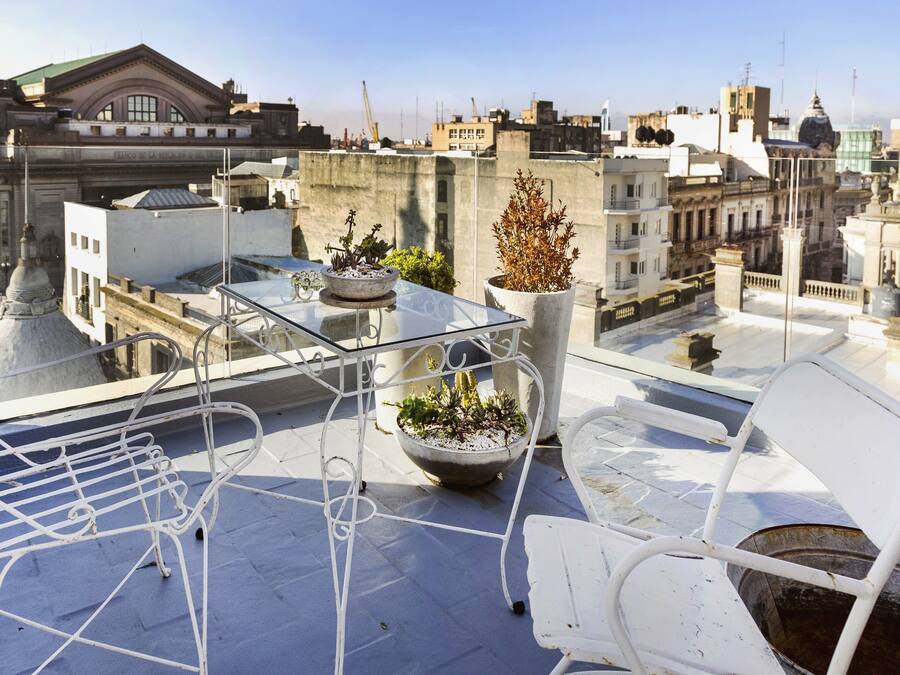 Terraza en la azotea con una mesa de vidrio, dos plantas en macetas y una silla de metal blanco.