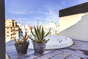 Dos plantas en macetas sobre una terraza de madera con un jacuzzi y una vista panorámica de la ciudad al fondo.
