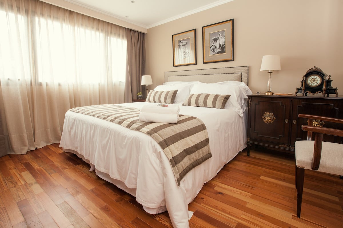 Een hotelkamer met een groot bed, een houten vloer, een nachtkastje met een lamp en een dressoir met een klok.
