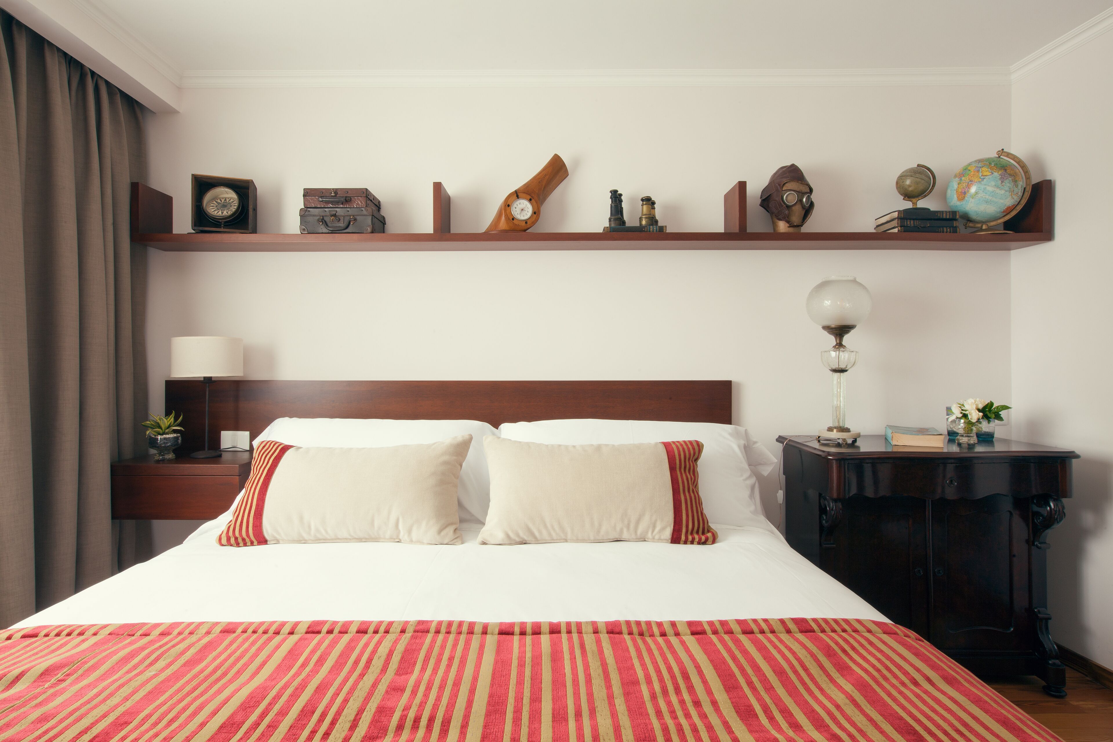 Una habitación de hotel con una cama grande, una mesita de noche con una lámpara, un cabecero de madera, un estante con objetos decorativos y una mesita auxiliar de madera oscura.