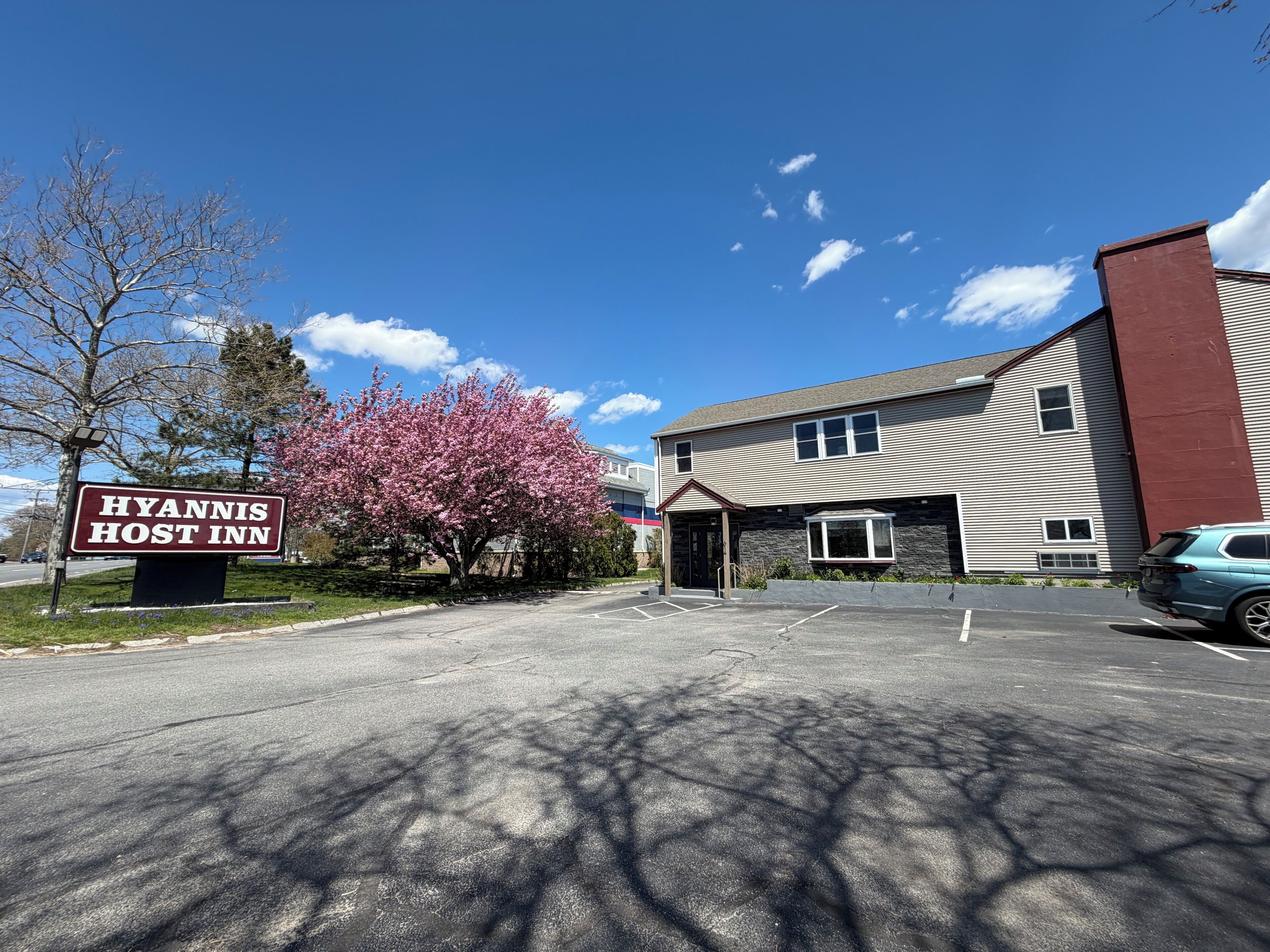 Un edificio con un cartello che recita 'Hyannis Host Inn', un parcheggio e un albero con fiori rosa.