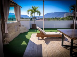 Una terraza en la azotea con cubierta de madera, una silla de descanso, una mesa y vistas a montañas y edificios.