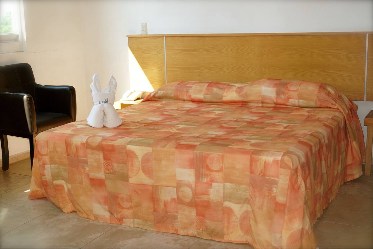 Una habitación de hotel con cabecera de madera, cama con colcha estampada, un peluche blanco sobre la cama y una silla de cuero negro.