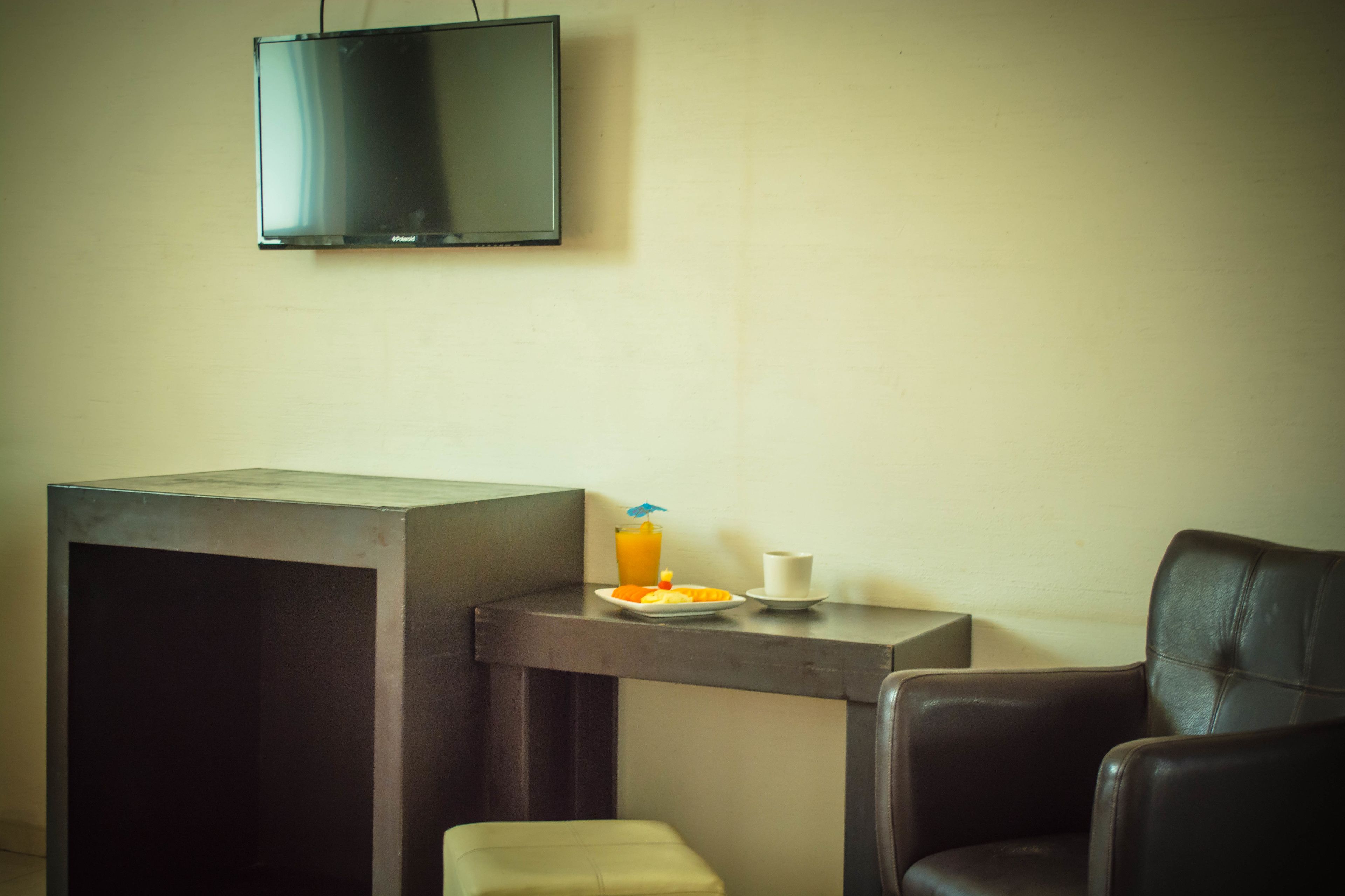Una habitación pequeña con un televisor en la pared, una mesa con un plato de comida y una bebida, una silla y un taburete.