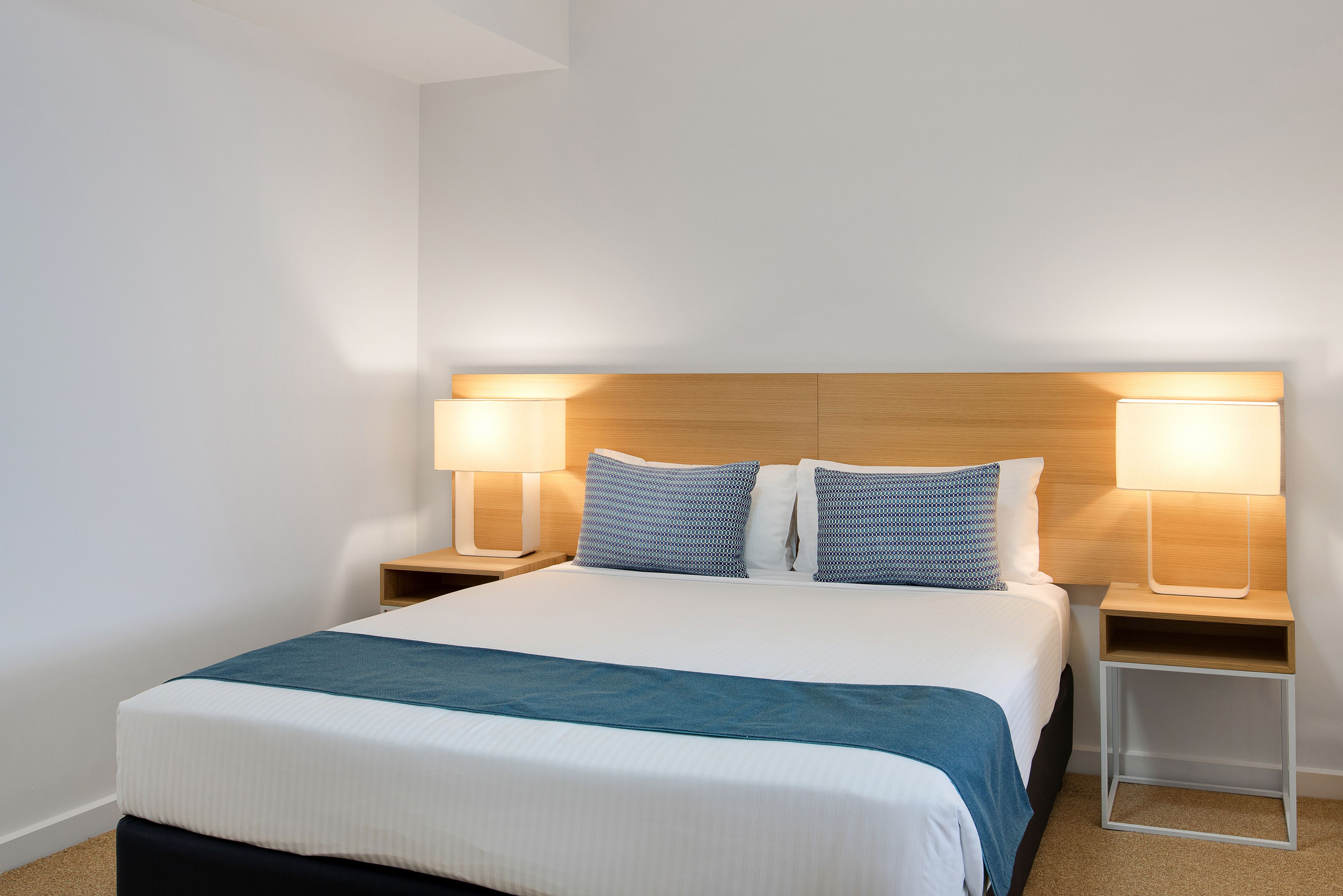 Ein ordentlich bezogenes Bett mit blauer Tagesdecke, zwei Nachttischlampen und einem hölzernen Kopfteil.