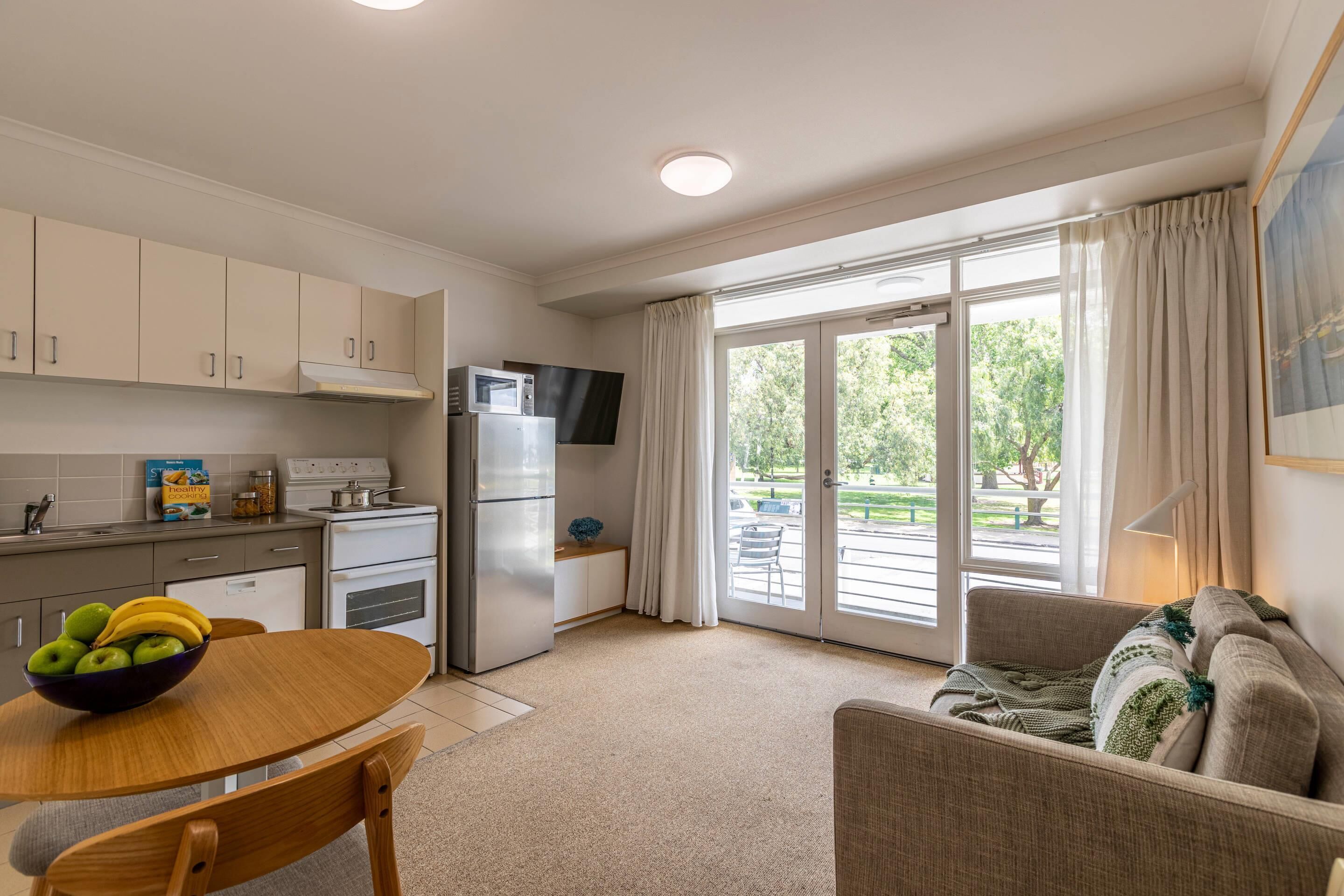 A modern kitchen with white cabinets, a microwave, and a refrigerator. There is a dining table with a bowl of fruit and a chair. The room has a large sliding glass door leading to an outdoor area with a view of trees.