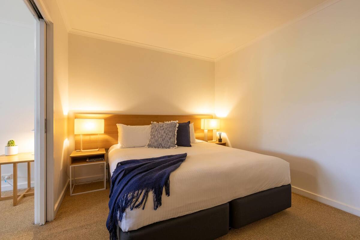 A neatly made bed with a dark headboard, two bedside lamps, a small bedside table with a plant, and a dark-colored blanket.