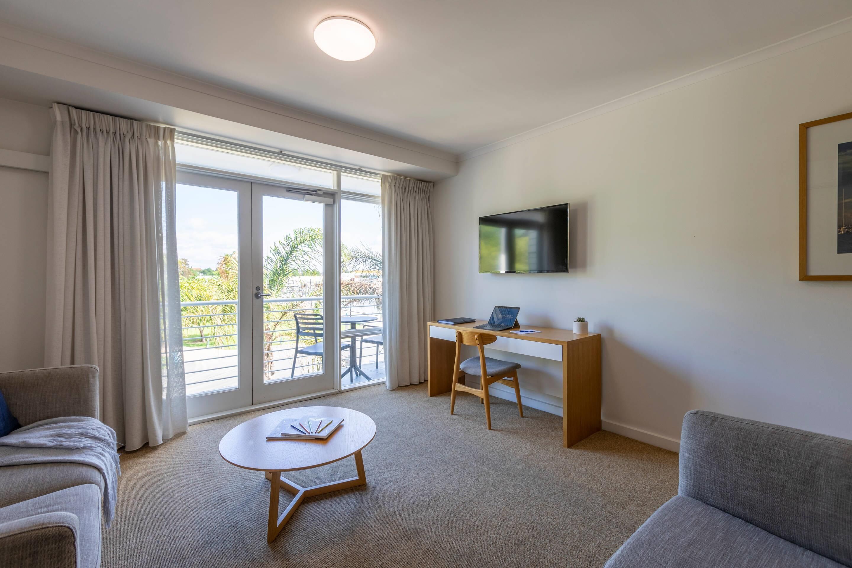Ein modernes Wohnzimmer mit einer Couch, einem Couchtisch, einem Schreibtisch mit Stuhl, einem an der Wand befestigten Fernseher und großen Fenstern mit Vorhängen.