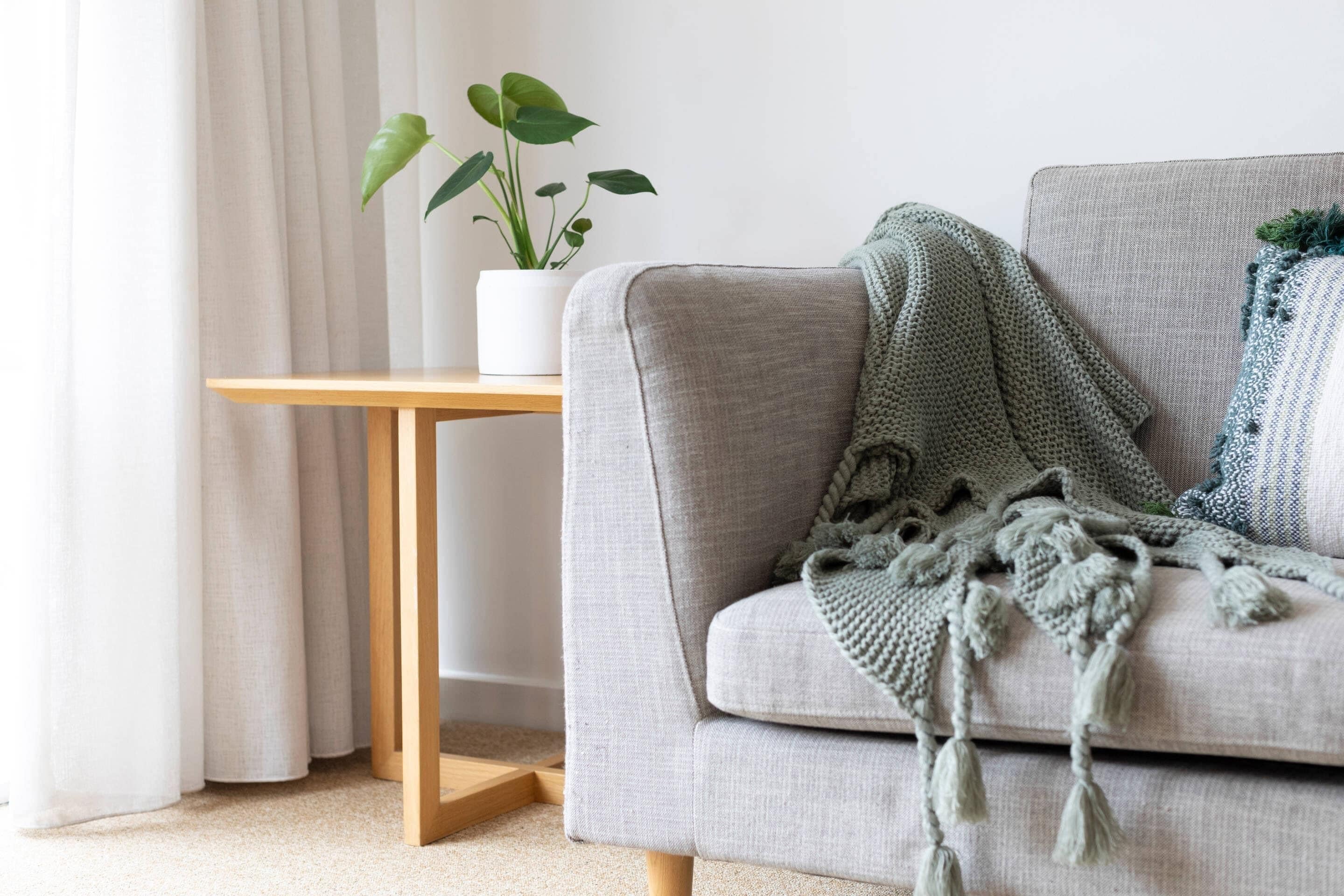 Un sofá gris con un plaid de punto verde, una planta blanca en maceta y una mesita de madera.