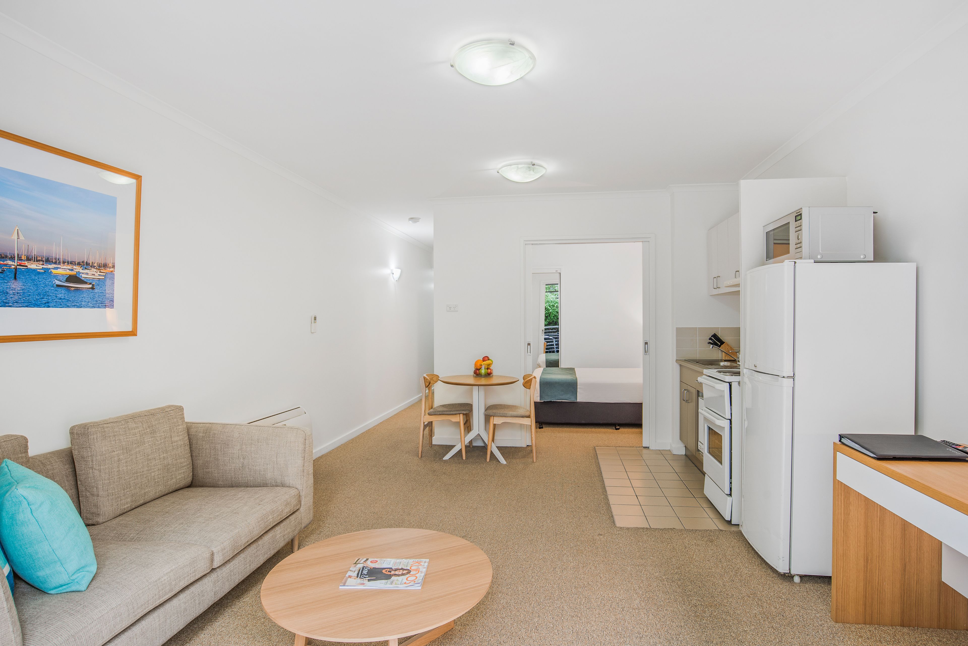 Una sala de estar moderna y bien iluminada con un sofá, una mesa de centro y un cuadro en la pared. Se observa un área de cocina con electrodomésticos blancos y una mesa de comedor.
