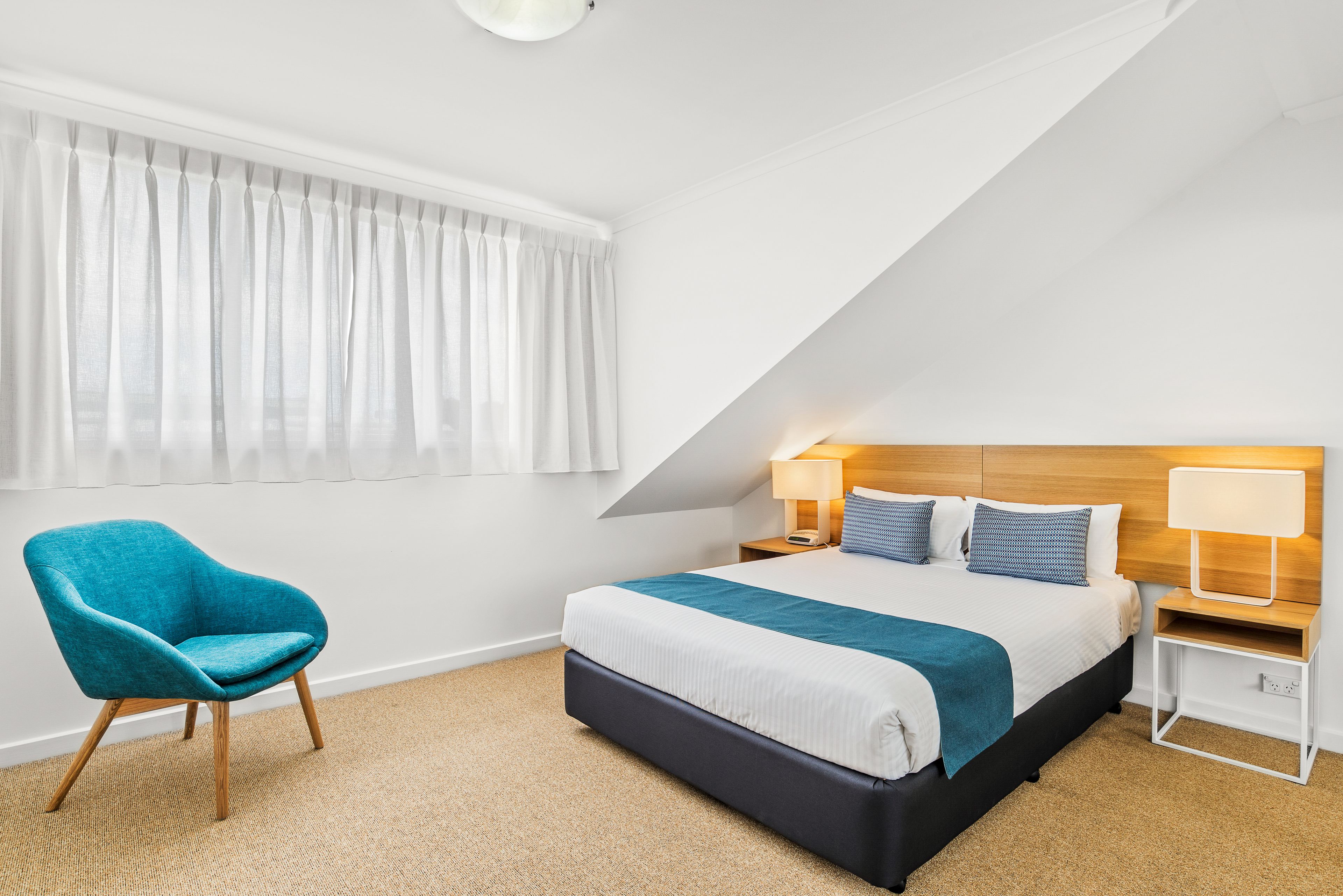 Una habitación de hotel moderna con una cama grande, un sillón verde azulado y un cabecero de madera.