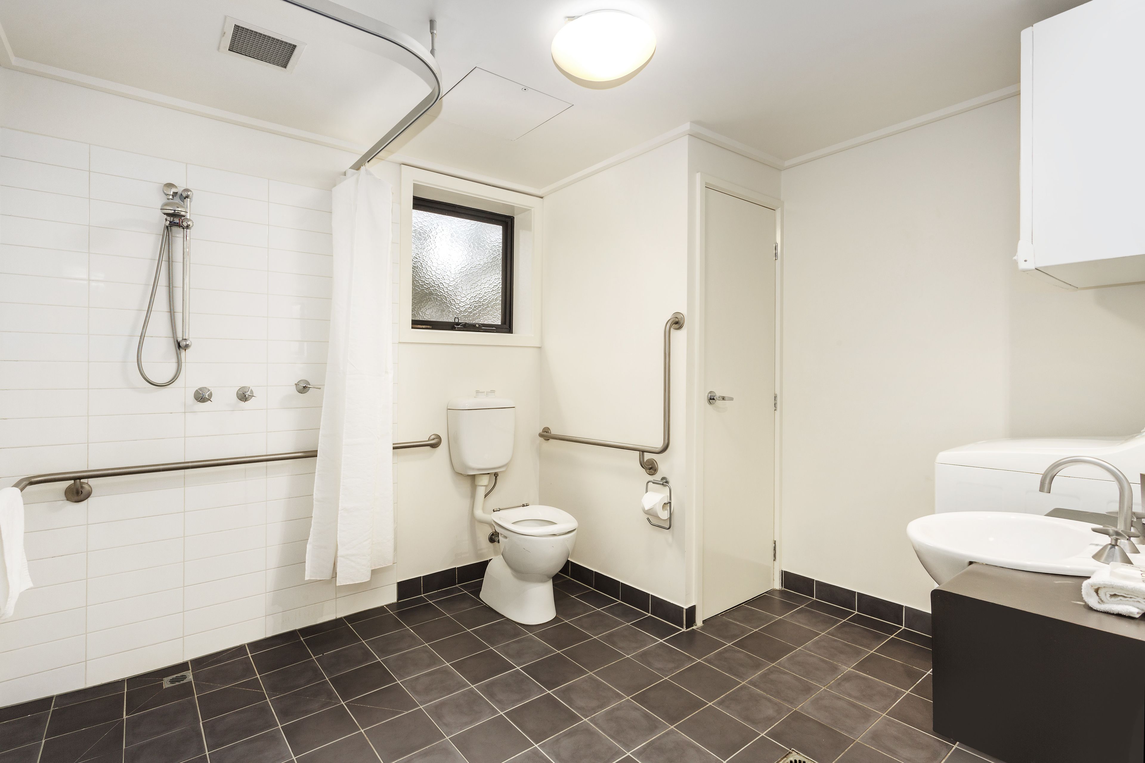 A modern bathroom with a white toilet, a shower with grab bars, a window, and a sink with a mirror.