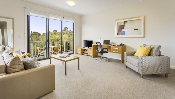 A modern living room with a sofa, a coffee table, a desk with a computer, and a TV. There is a balcony with a view of greenery. - Quest Frankston (Frankston)
