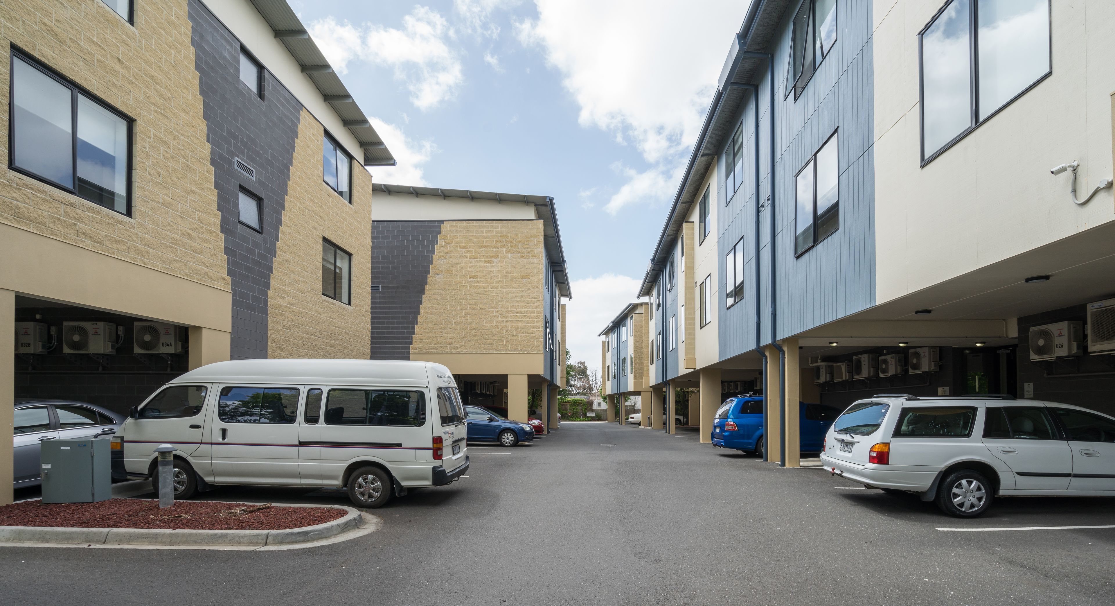 A street view of a residential area with multi-story buildings, parked cars, and a clear sky.