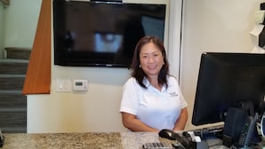 A person standing at a reception desk with a television mounted on the wall above, a thermostat, and a computer monitor to the right. - Marin Lodge (San Rafael)