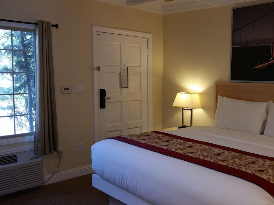 A hotel room with a bed, a nightstand, a lamp, a door, and a window with curtains.