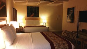 A hotel room with two beds, a wooden headboard, a nightstand with a lamp, a painting on the wall, a small table with a chair, and a digital clock.