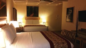 A hotel room with two beds, a wooden headboard, a nightstand with a lamp, a painting on the wall, a small table with a chair, and a digital clock.