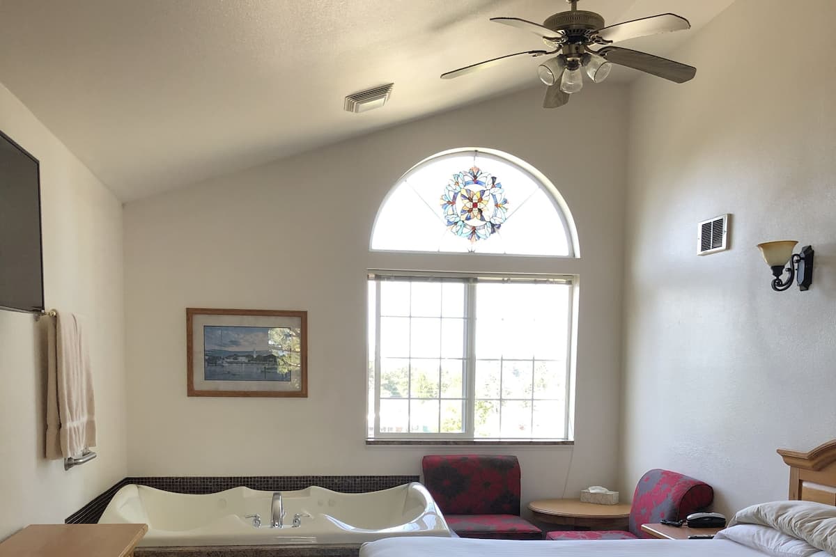A hotel room with a large bed, a bathtub, a vanity with a sink, a ceiling fan, a window with a stained glass design, and a framed picture on the wall.