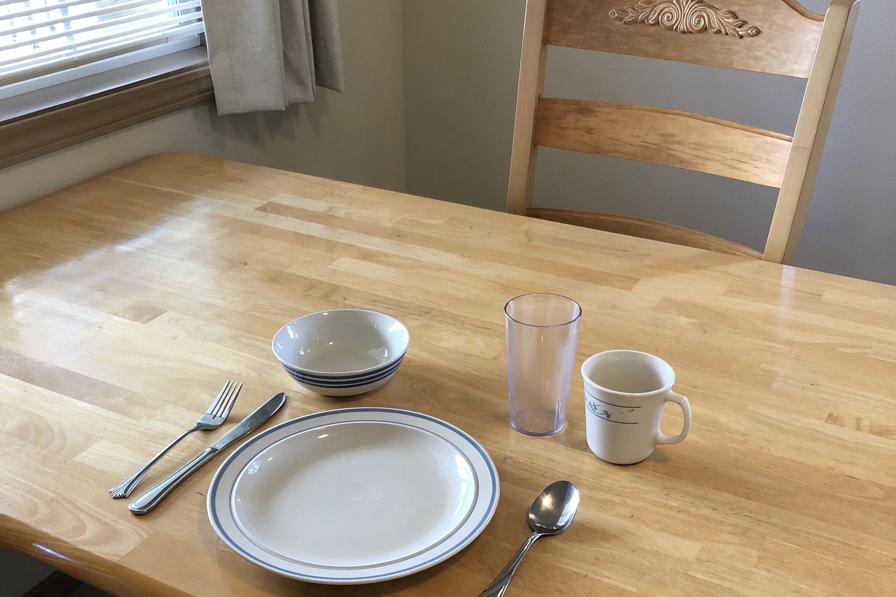 A wooden dining table with a plate, bowl, glass, mug, fork, knife, and spoon.