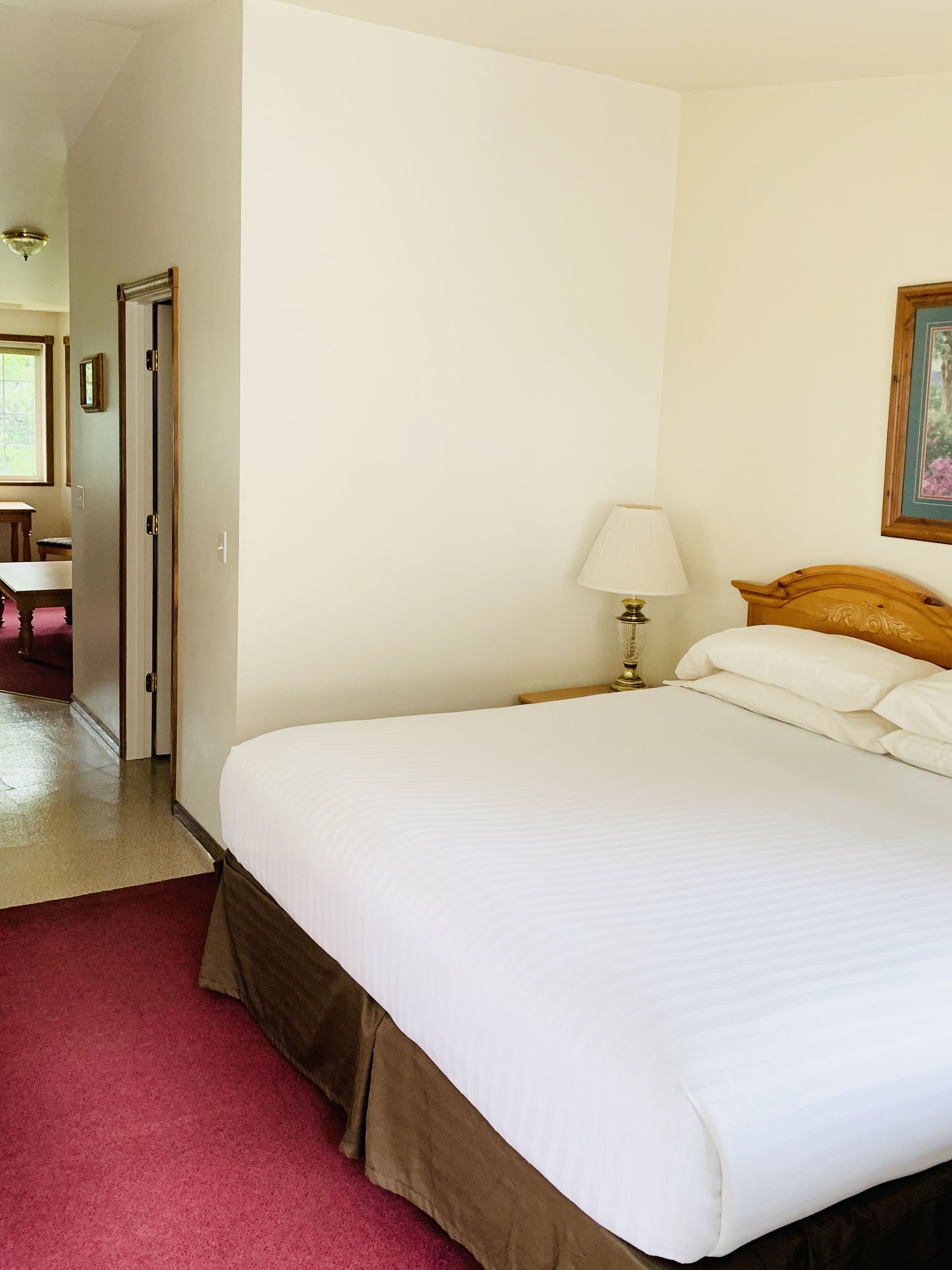 A hotel room with a large bed, a bedside table with a lamp, a door leading to another room, and a framed picture on the wall.