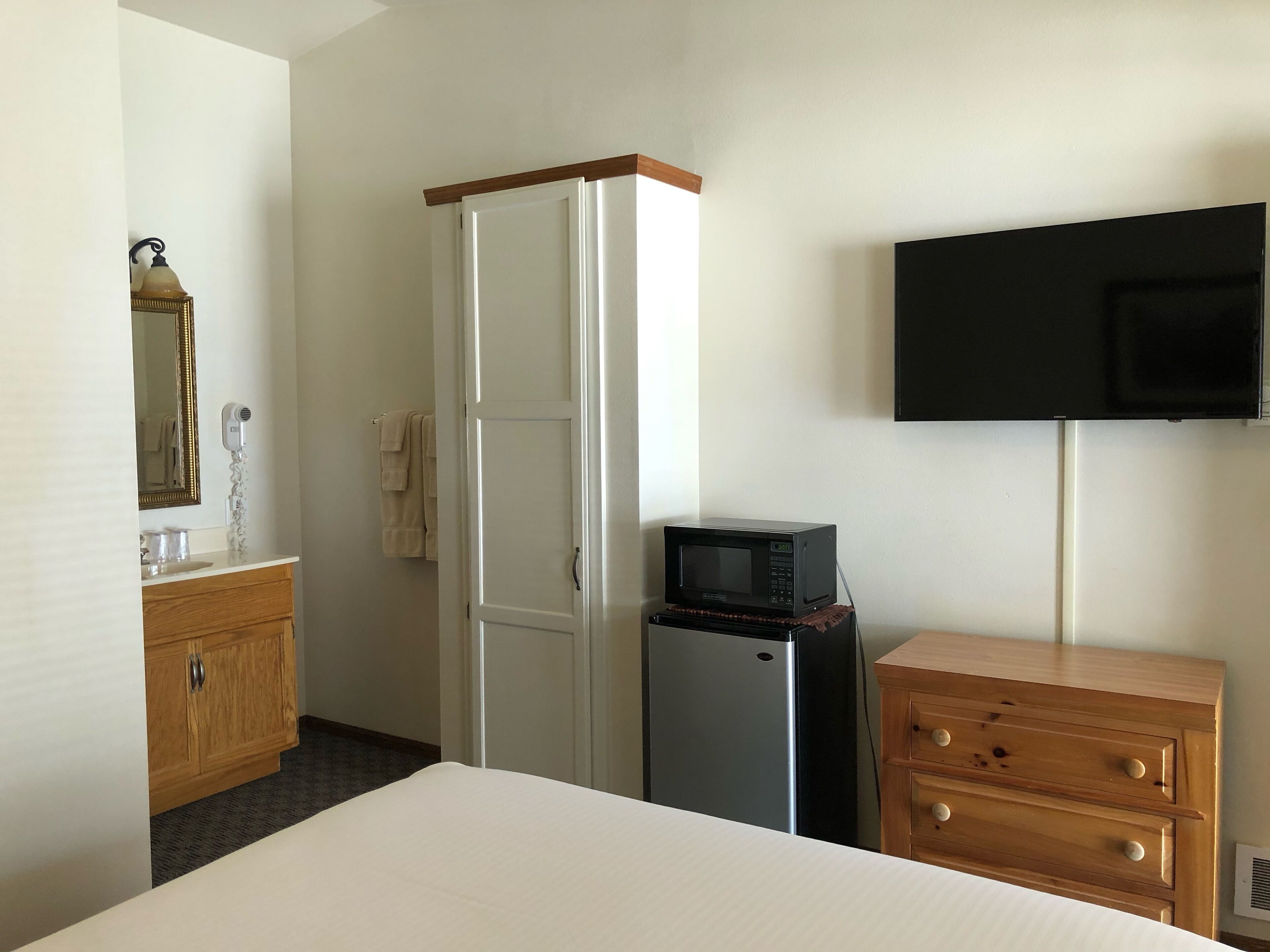 Habitación de hotel con cama, cómoda de madera, microondas, televisión, armario y tocador con espejo y secador de pelo.