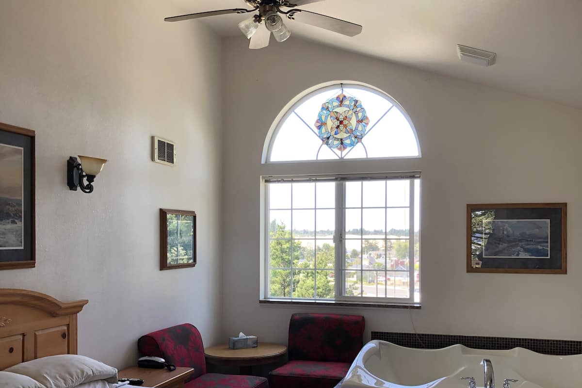 A hotel room with a bed, a seating area with a table, a large window with a stained glass design, and a bathtub.