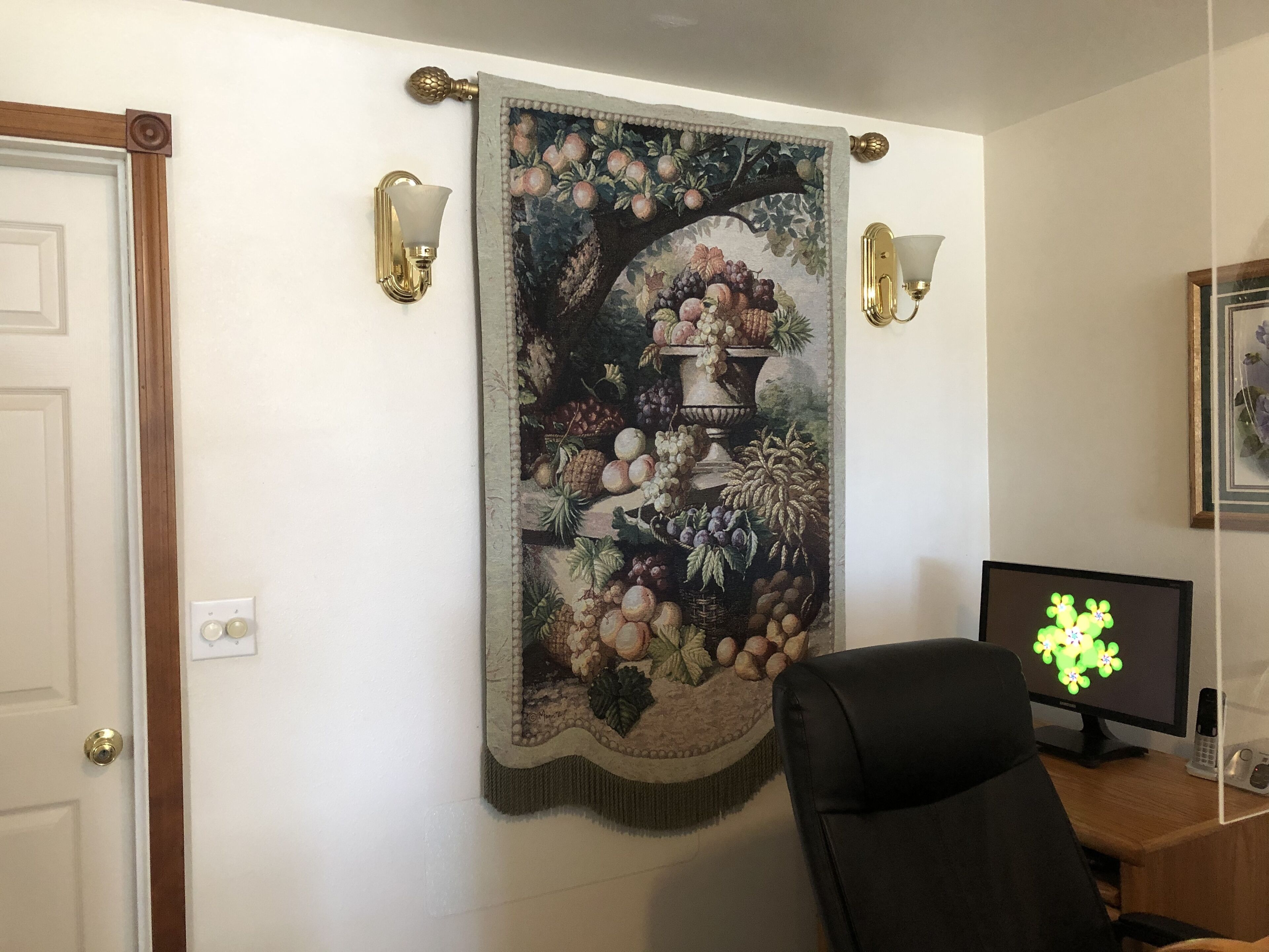 Una stanza con un grande arazzo raffigurante frutta e un vaso, due lampade a parete, un monitor per computer e una porta.