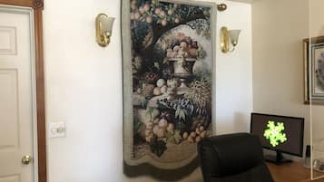 A room with a large tapestry depicting fruits and a vase, two wall-mounted lamps, a computer monitor, and a door.