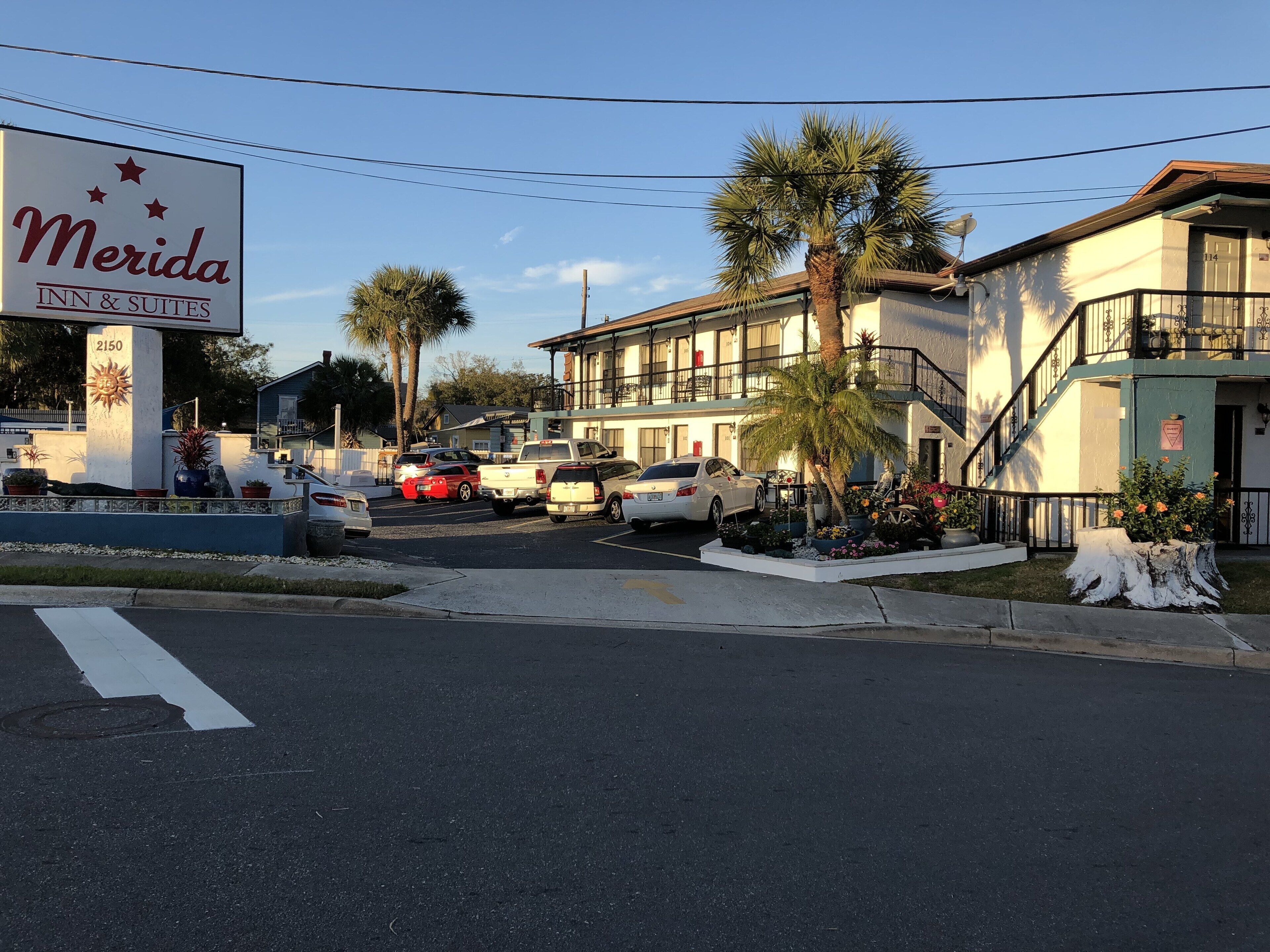 A hotel named 'Merida Inn & Suites' with a sign featuring a sun and stars. There are parked cars, a palm tree, and a building with a balcony.