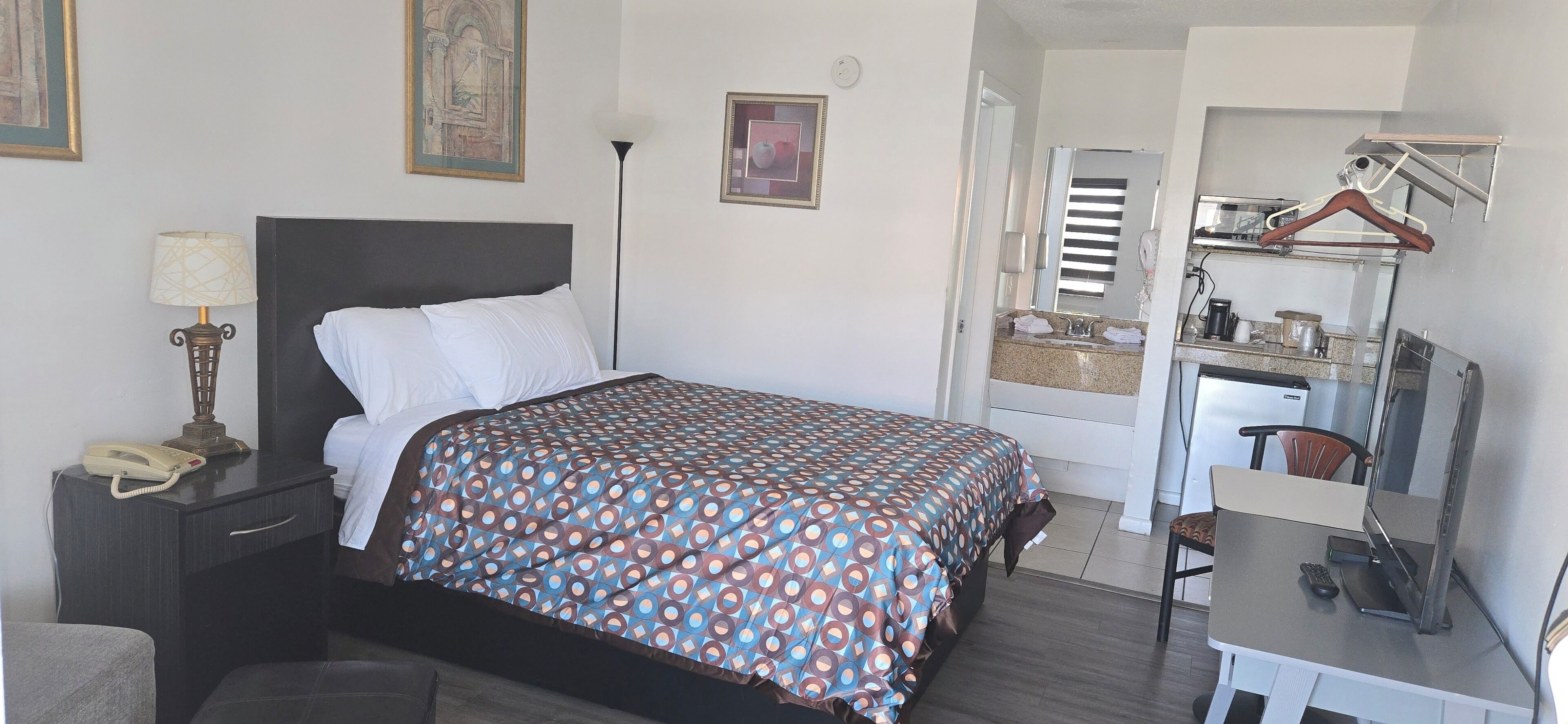 A hotel room with a bed, bedside table with a lamp, a desk with a chair, a mirror, and a hanging coat rack.