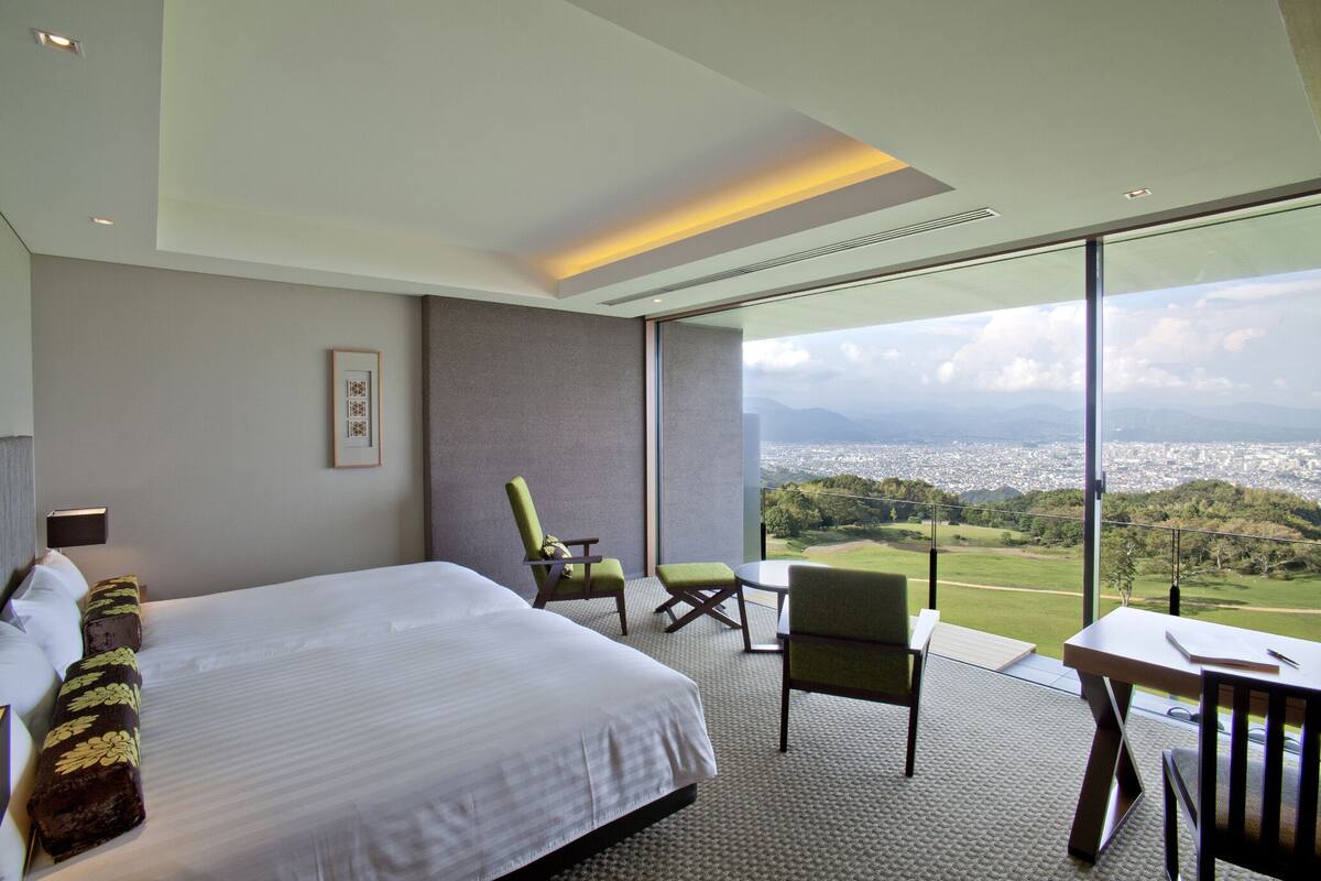 A modern hotel room with a large bed, a seating area with chairs, and a view of a cityscape and mountains through large windows.