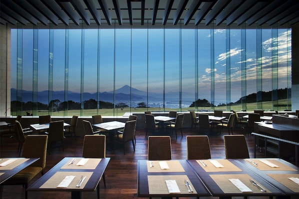 A restaurant with large windows offering a view of a mountainous landscape. Tables are set with placemats and cutlery.
