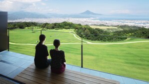 Dos personas sentadas en un balcón con vistas a un campo de golf, un paisaje urbano y una montaña al fondo.