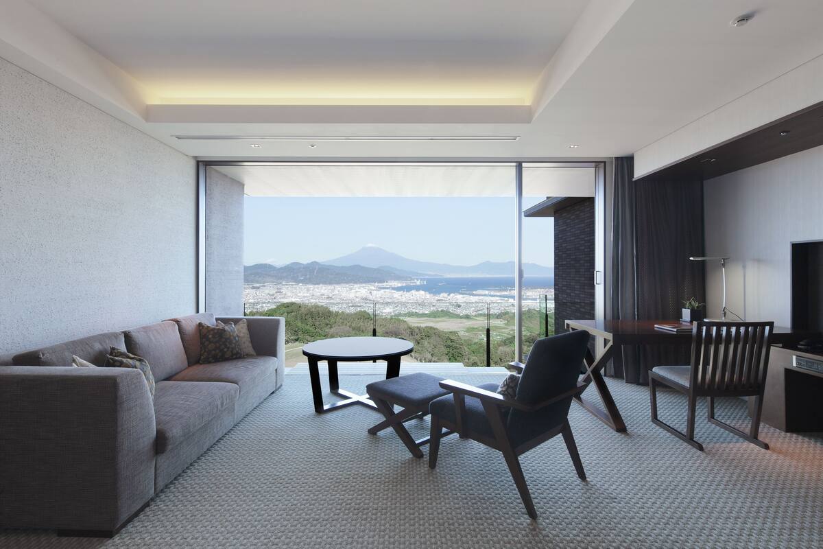 A modern living room with a sofa, chairs, a small table, and a large window offering a city and mountain view.